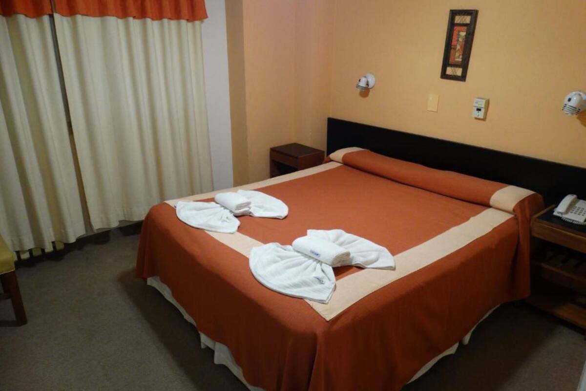 Quarto de hotel com cama bem arrumada, mesa de cabeceira com telefone e luminária fixada na parede.