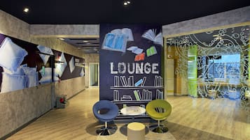 A modern lounge area with a large wall displaying 'Lounge' and images of books, a small table with magazines, and two colorful chairs.