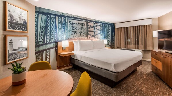 Habitación de hotel con cama, mesitas de noche, televisor, escritorio y sillas. En la pared hay cuadros enmarcados y un mural grande de un puente.