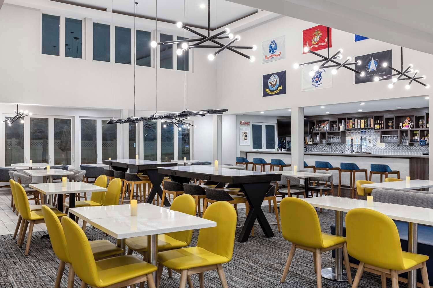 A modern dining area with multiple tables and chairs, a bar area with shelves, and flags on the walls.