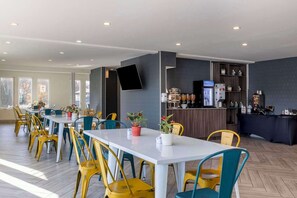 Un comedor moderno con varias mesas y sillas, una barra con máquina de café, y un televisor de pantalla plana en la pared.