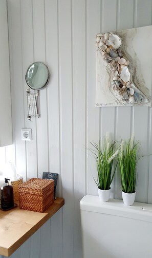 A bathroom with a wooden shelf holding toiletries, a mirror, and a decorative piece above the toilet.