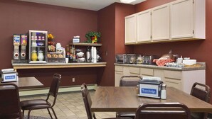 A dining area with a wooden table and chairs, a coffee machine, a refrigerator stocked with beverages, and a small kitchen area with cabinets and a microwave. - Travelodge by Wyndham Motel of St Cloud (St Cloud)