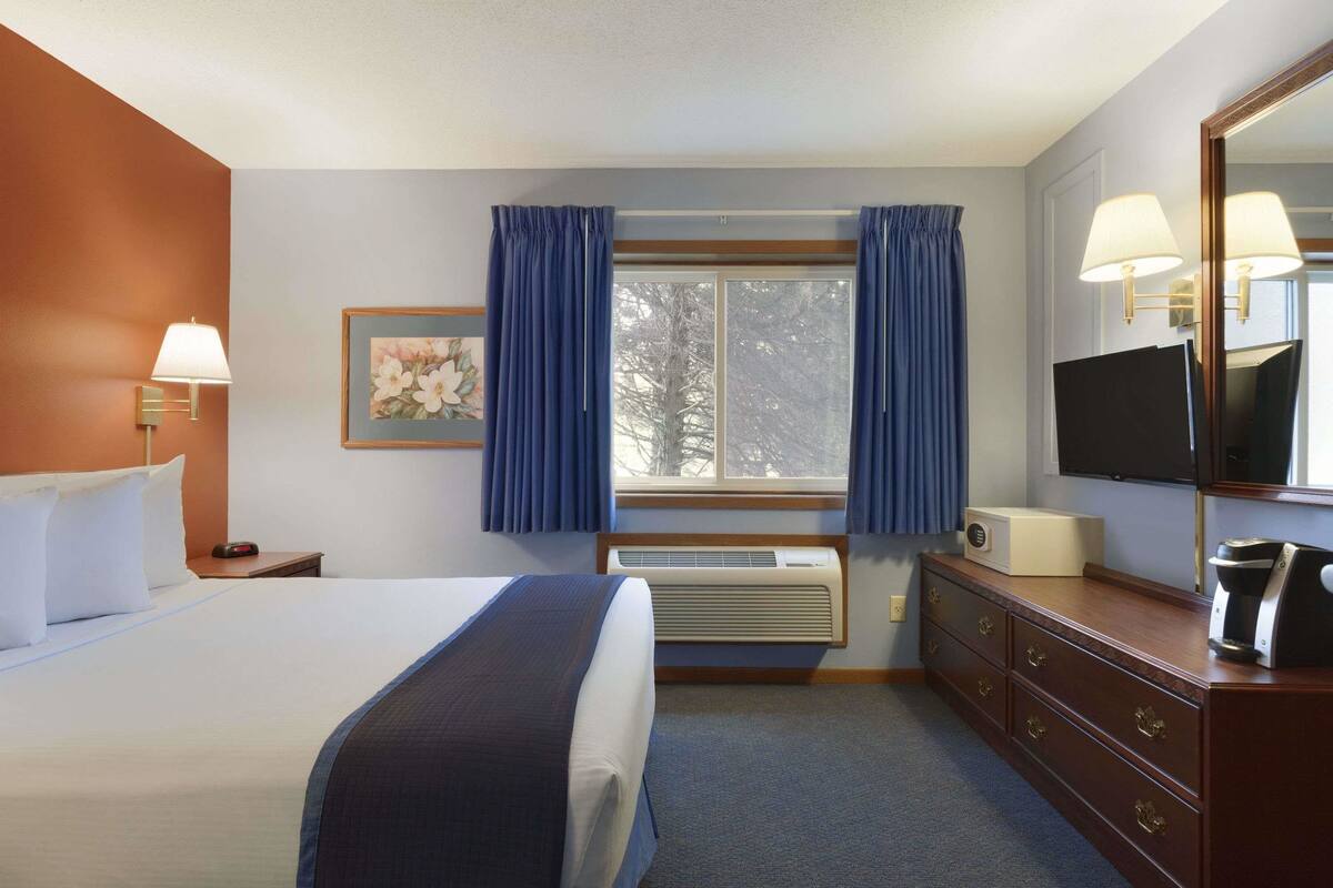 A hotel room with a bed, a dresser, a television, a window with curtains, and a wall-mounted air conditioner.
