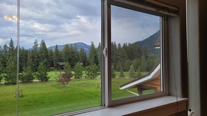 A room with a large window overlooking a green landscape with trees and a building with a sloped roof. - Mountain View Studio Apartment, Close to GNP (Columbia Falls)