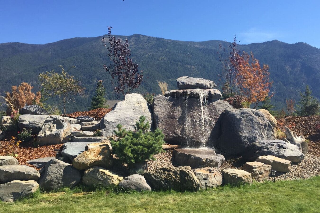 Une formation rocheuse naturelle avec une cascade, entourée d’un feuillage automnal et de montagnes en arrière-plan.