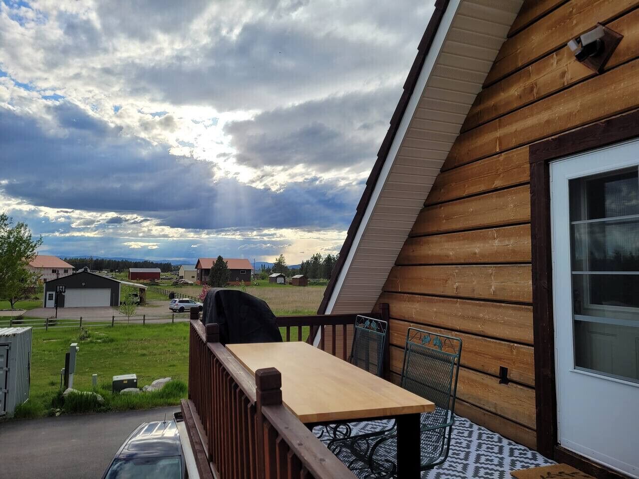 A wooden deck with a table and chairs, overlooking a grassy area with houses in the background.