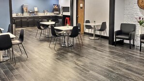 A modern dining area with tables and chairs, a kitchen in the background, and a brick wall. - Super 8 by Wyndham Georgetown (Georgetown)