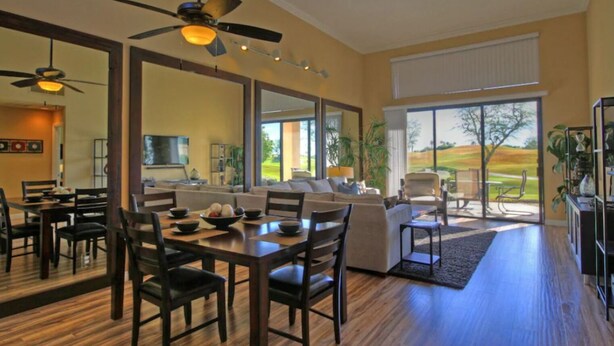 A spacious living area with a dining table, chairs, and a sofa. There are ceiling fans, large windows, and a view of a golf course. - Remodeled condo on PGA West's Stadium Golf Course (La Quinta)