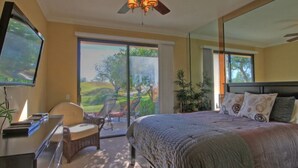 A bedroom with a bed, a television, a desk, and a chair. There is a sliding glass door leading to a balcony with chairs and a view of a green landscape. - Remodeled condo on PGA West's Stadium Golf Course (La Quinta)