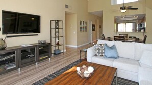 A living room with a flat-screen TV, a coffee table with decorative items, a white sofa with patterned cushions, and a dining area with chairs and a table. - Remodeled condo on PGA West's Stadium Golf Course (La Quinta)