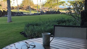 A table with a book and a mug, overlooking a well-maintained lawn with palm trees and a view of a building in the distance. - Secluded 2-Suite - Stocked Like you're at home. Ocean front pool, private lanai (Waikoloa Village)