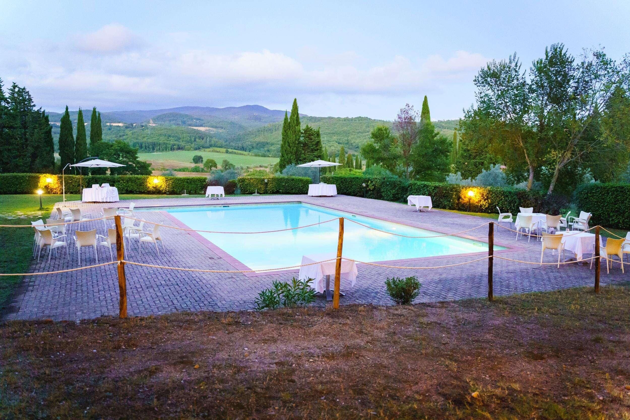 A rectangular pool with a paved deck, surrounded by white lounge chairs and umbrellas, set against a backdrop of rolling hills and trees.