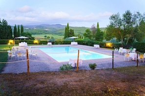 Una piscina rettangolare con bordo piastrellato, circondata da poltroncine da sole bianche e ombrelloni, con vista su dolci colline e alberi.