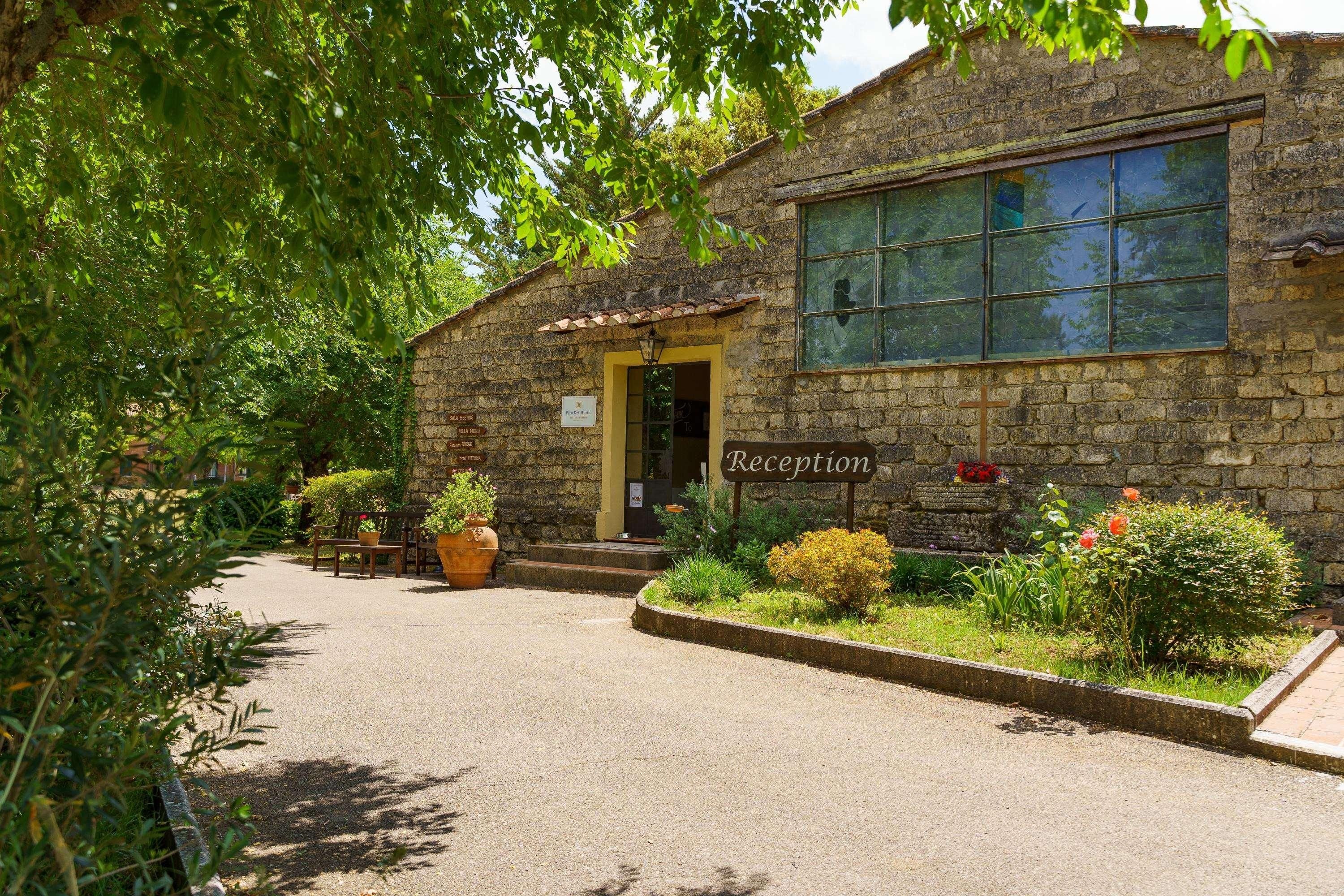 Un bâtiment en pierre avec une enseigne indiquant l’accueil, entouré de verdure et agrémenté de plantes en pot.