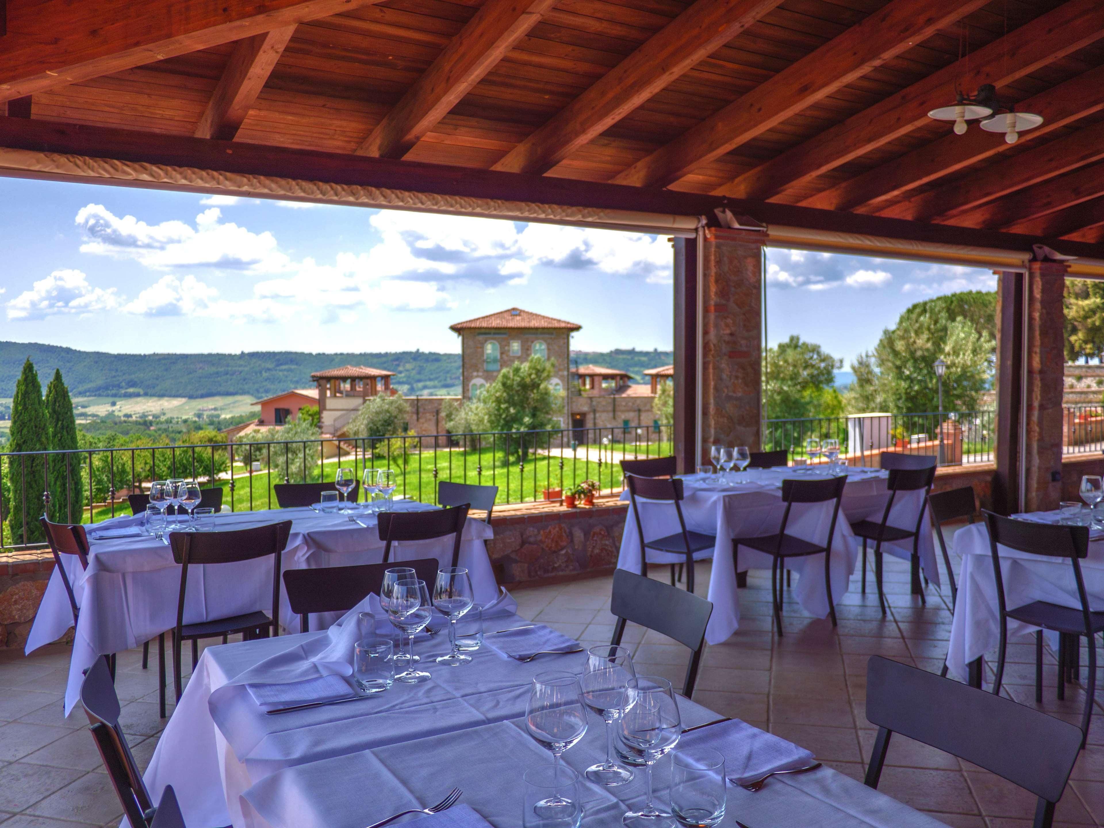 An outdoor dining area with tables set for two, overlooking a scenic landscape with buildings and trees.
