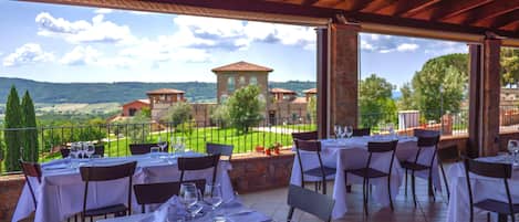 An outdoor dining area with tables set for two, overlooking a scenic landscape with buildings and trees.