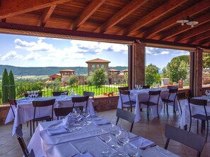 Un área de comedor al aire libre con mesas preparadas para dos, con vistas a un paisaje pintoresco con edificios y árboles.