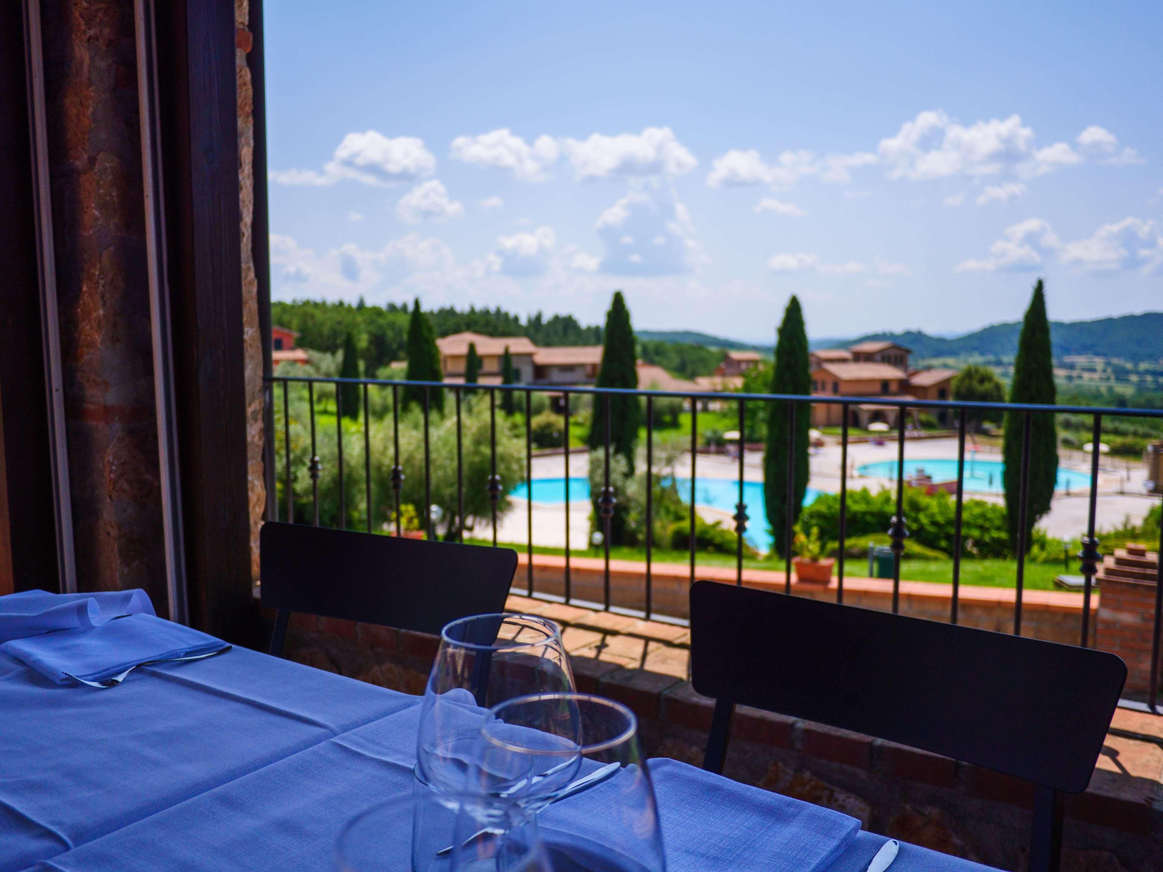A table with a blue tablecloth and two wine glasses on a balcony overlooking a swimming pool and greenery.