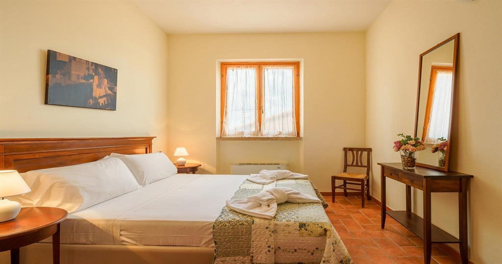 Habitación de hotel con cama, mesita de noche con lámpara, silla, ventana con cortinas, espejo y un cuadro en la pared.