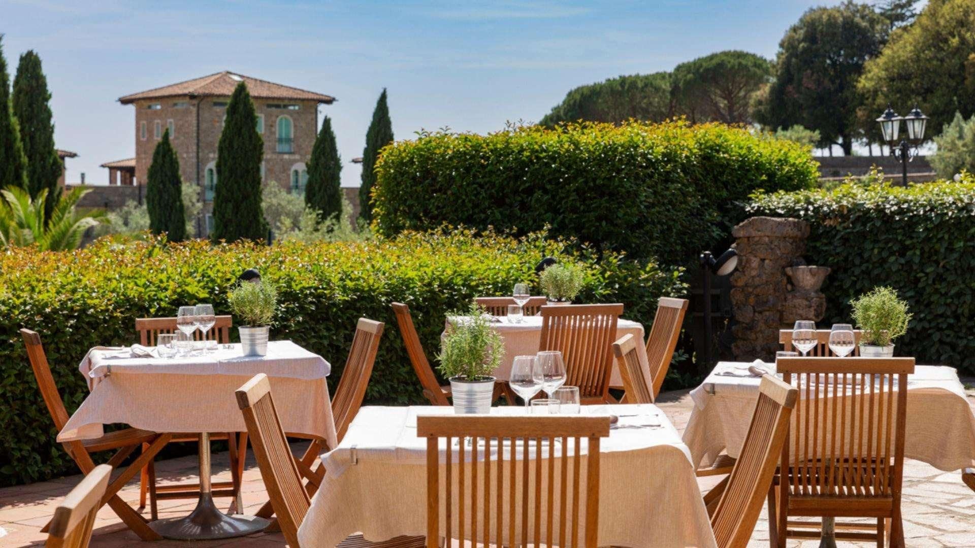 An outdoor dining area with tables and chairs, surrounded by greenery and a stone wall.
