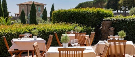 An outdoor dining area with tables and chairs, surrounded by greenery and a stone wall.