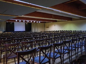 Un'ampia sala con file di sedie di legno e un palco con uno schermo.