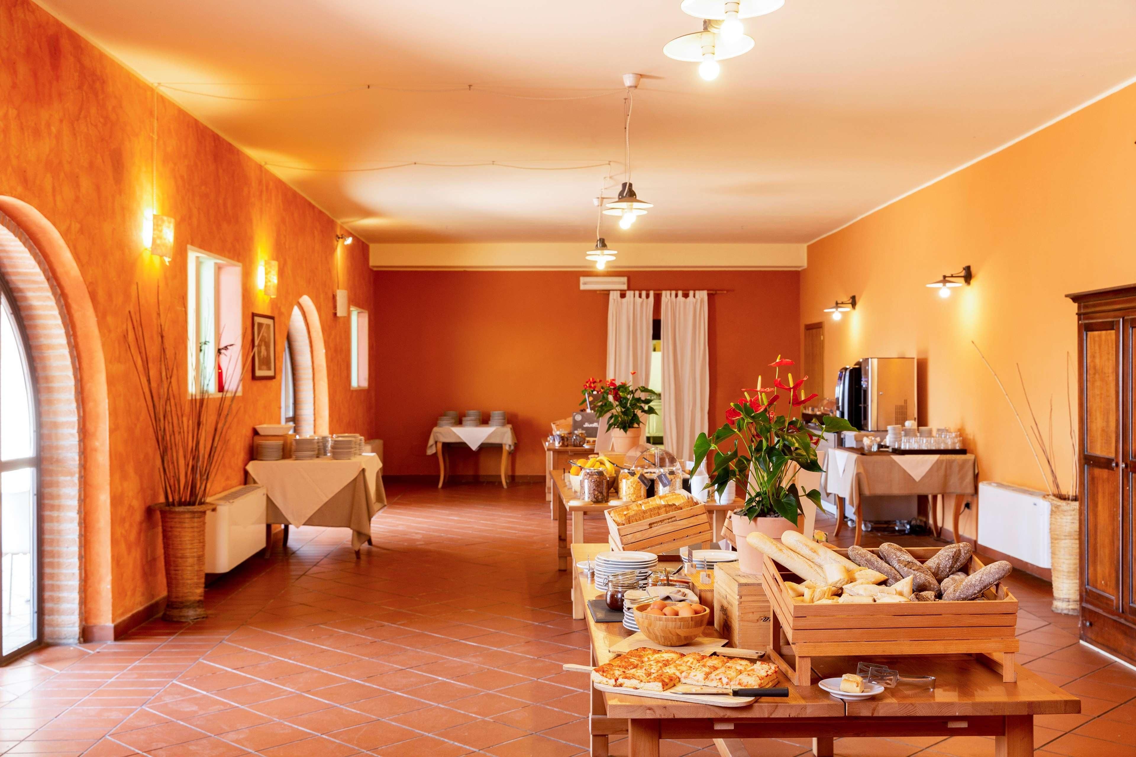 Un espace repas avec un buffet proposant du pain, des viennoiseries et un coin dédié aux pizzas. On y trouve des tables et des chaises, et les murs de la pièce sont peints dans une couleur orange chaleureuse.