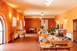 Un comedor con mesas y sillas, y una barra de desayuno tipo buffet que incluye pan, bollería y una estación de pizza. Las paredes son de un cálido color naranja.