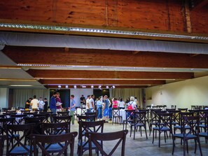 Un espacio interior amplio con varias mesas y sillas, vigas de madera en el techo y un escenario con iluminación.