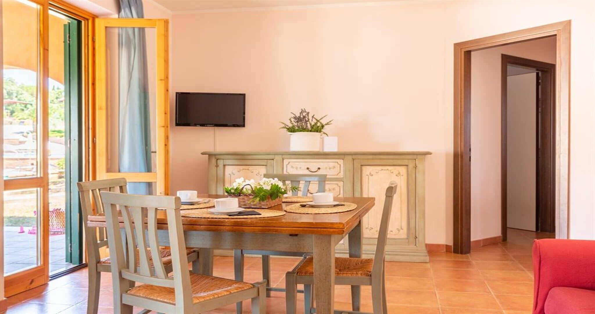 Un comedor con una mesa de madera, cuatro sillas, un televisor montado en la pared y un mueble con cajones.