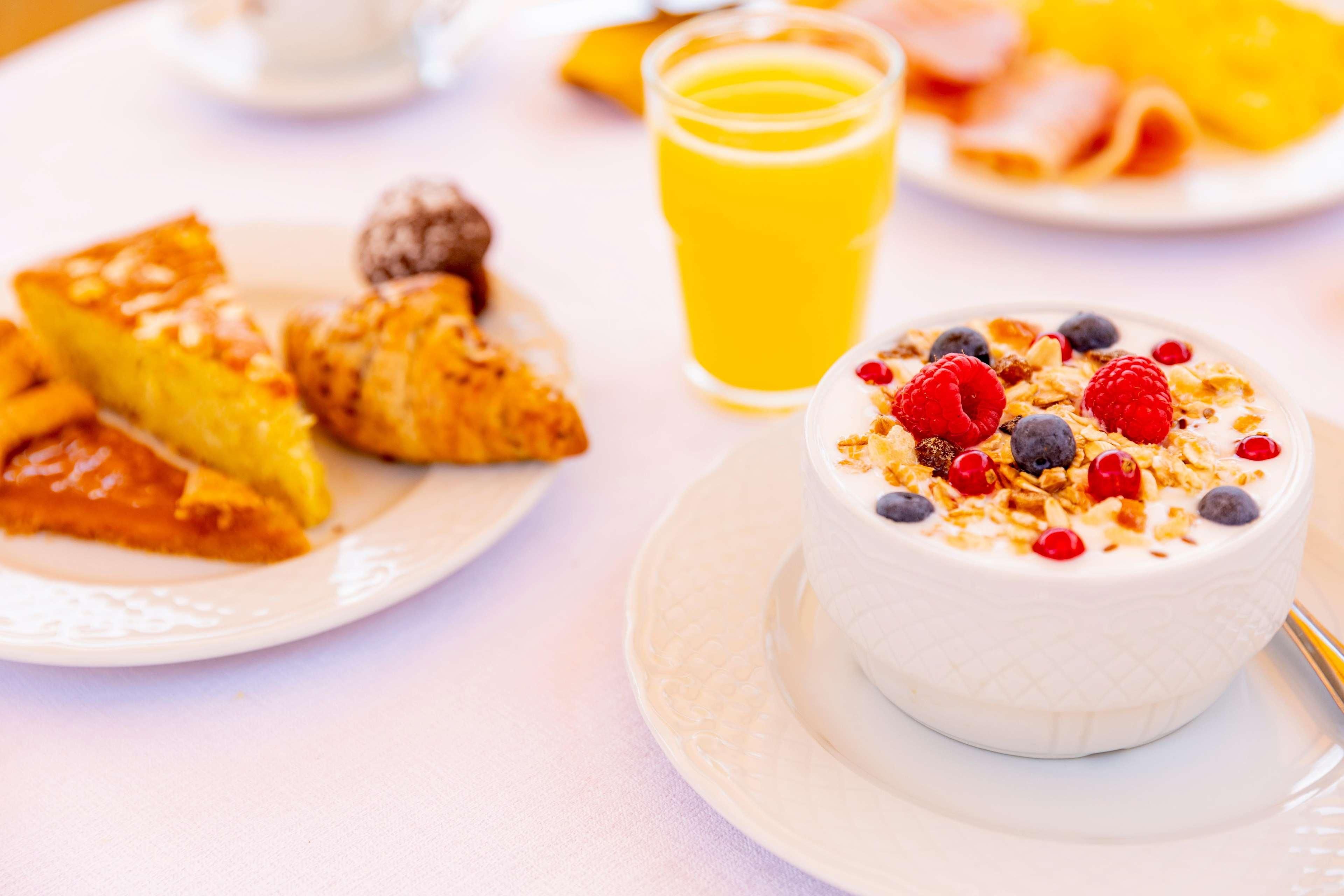 A breakfast setting with a bowl of yogurt topped with berries and granola, a glass of orange juice, and a plate with croissants and a slice of cake.