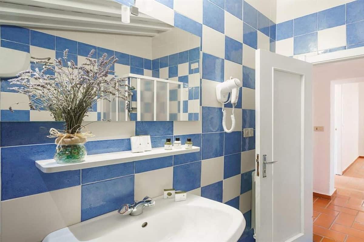 A bathroom with a white sink, blue and white tiles, a vase with dried flowers, and a hairdryer on the wall.