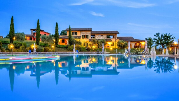 Una gran piscina bien iluminada con reflejos nítidos, rodeada por un jardín ajardinado y un edificio alto de varios pisos con terrazas al aire libre.