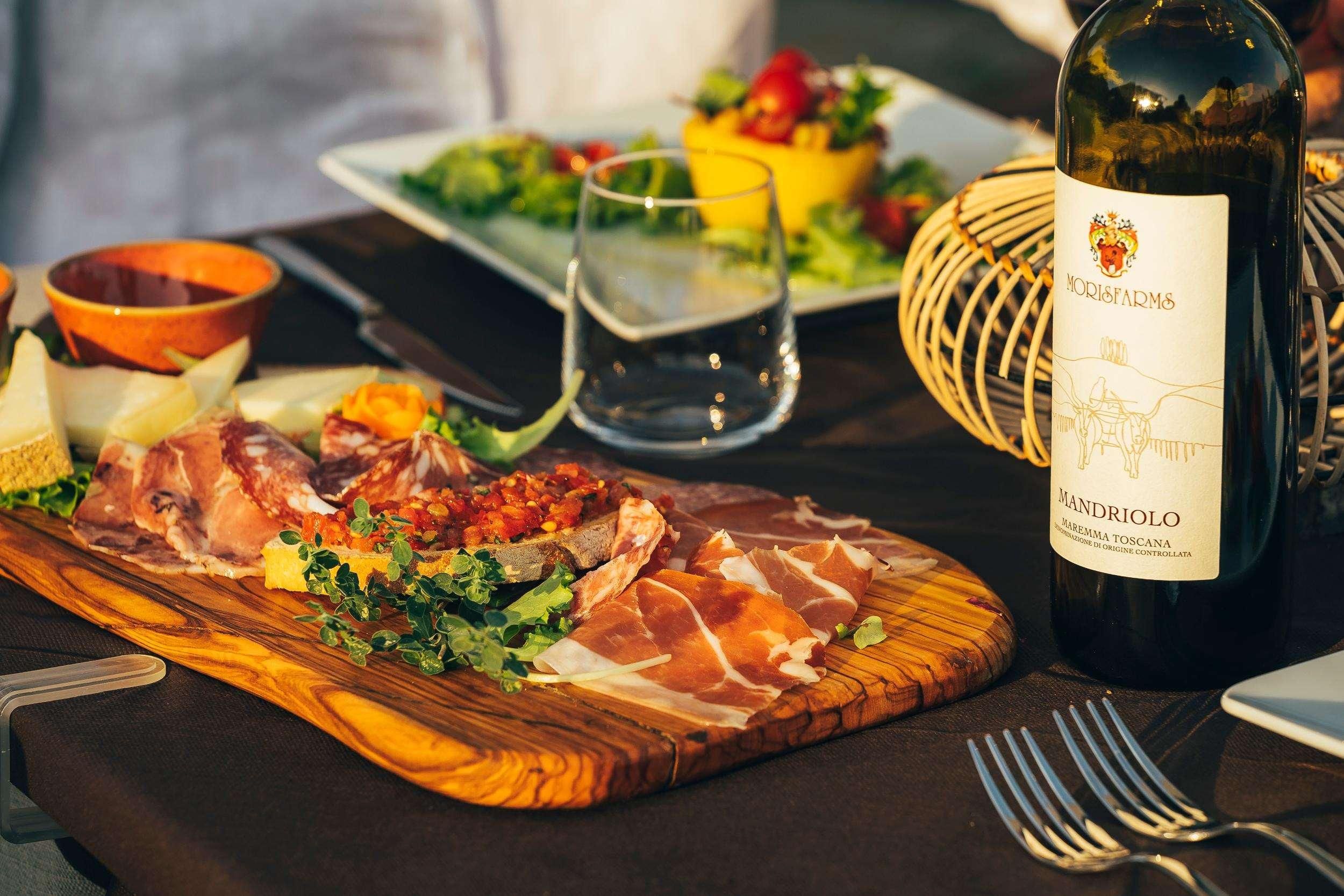 A wooden board with prosciutto, cheese, and a salad, a bottle of wine, and a glass of water on a table.