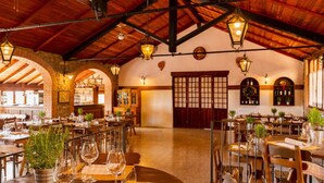 Un restaurante con vigas de madera, arcos de ladrillo y faroles colgantes. Las mesas están dispuestas con copas de vino y macetas.