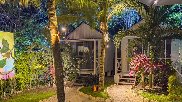 A well-lit garden with a gravel path, palm trees, and a wooden deck leading to a building with large windows. - Zensus Eco Boutique Hotel (Bacalar)