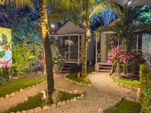 A well-lit garden with a gravel path, palm trees, and a wooden deck leading to a building with large windows. - Zensus Eco Boutique Hotel (Bacalar)