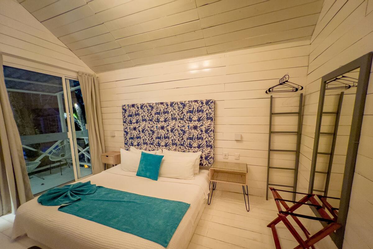 Un dormitorio con cama, cabecera estampada, mesita de noche, escalera de madera y ventana con cortinas.
