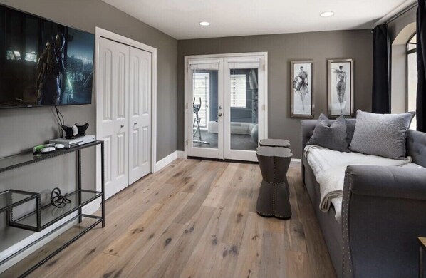 A modern living room with a grey sofa, a coffee table, and a TV mounted on the wall. There is a sliding glass door leading to a balcony.