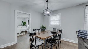 Un coin repas avec une table en bois et des chaises noires, un luminaire suspendu et une fenêtre avec des stores.