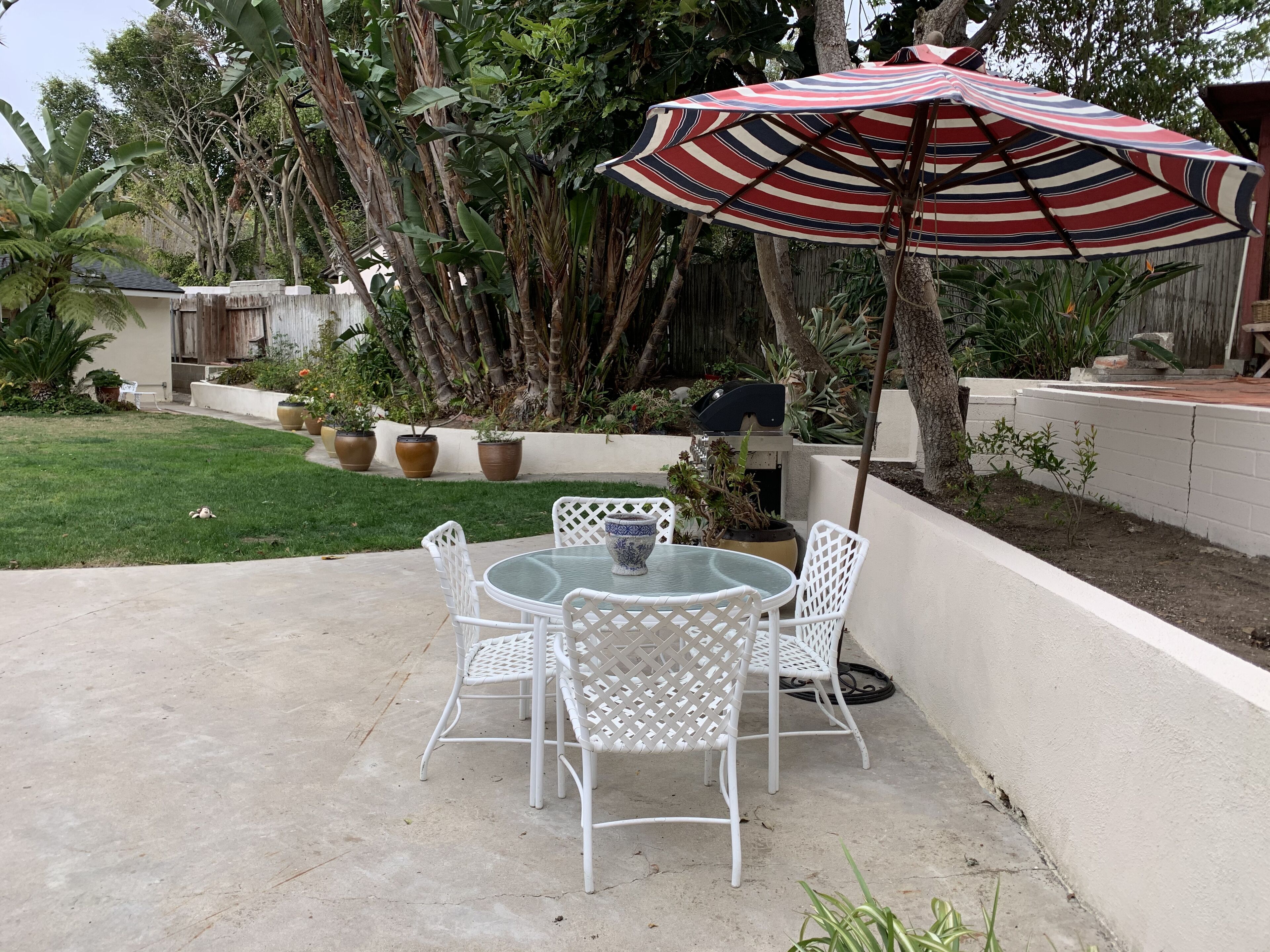 Eine Terrasse mit rundem Tisch und weißen Stühlen, umgeben von Blumentöpfen und einem gestreiften Sonnenschirm.