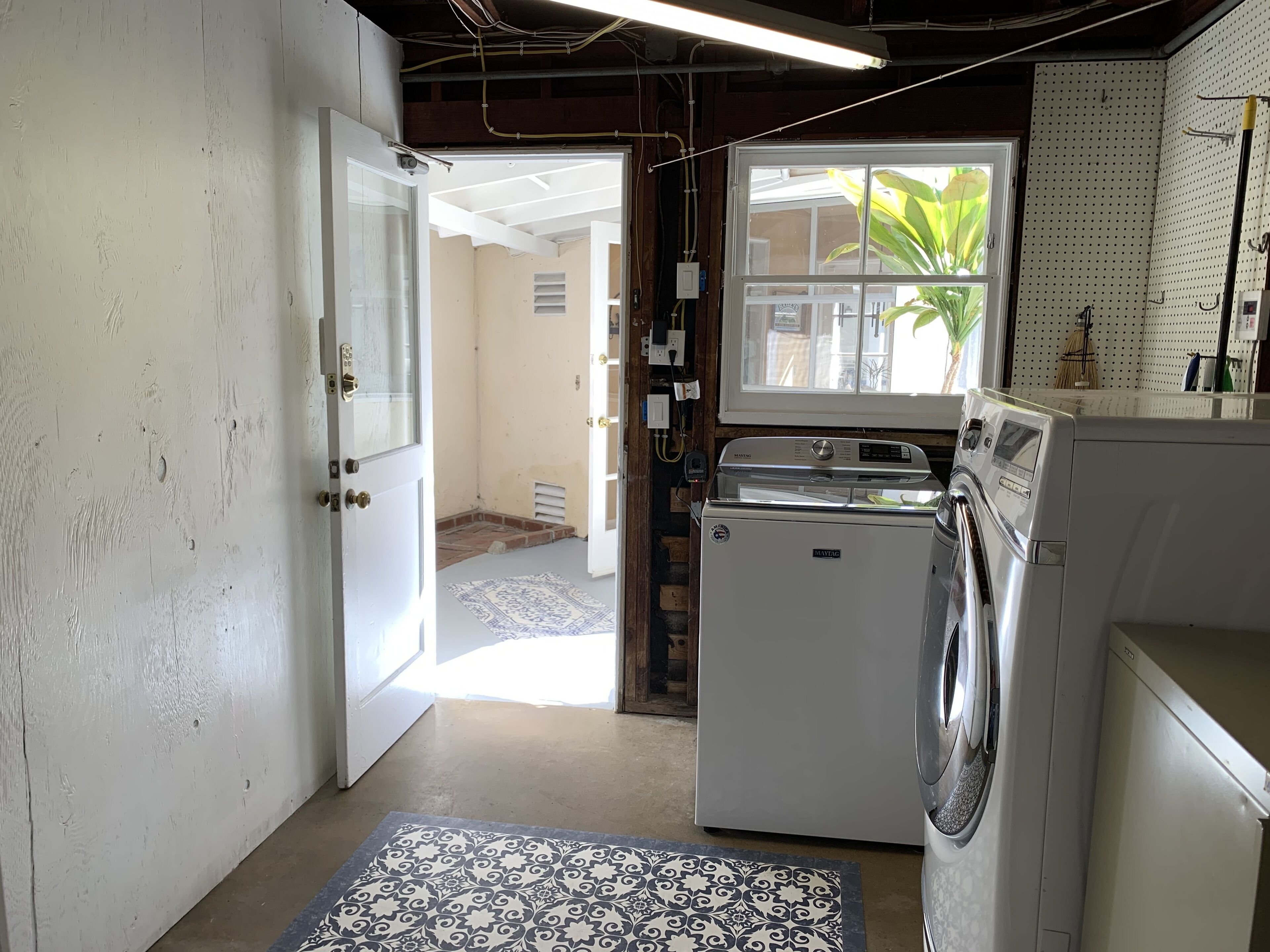 Une buanderie équipée d’un lave-linge et d’un sèche-linge, avec un tapis à motifs et une fenêtre agrémentée d’une plante.
