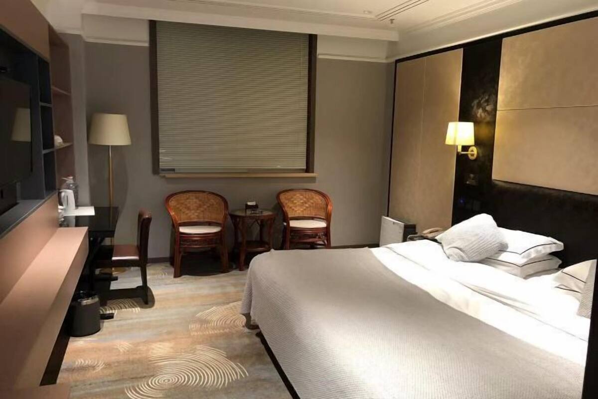 A hotel room with a bed, two chairs, a small table, a lamp, and a large window with blinds.