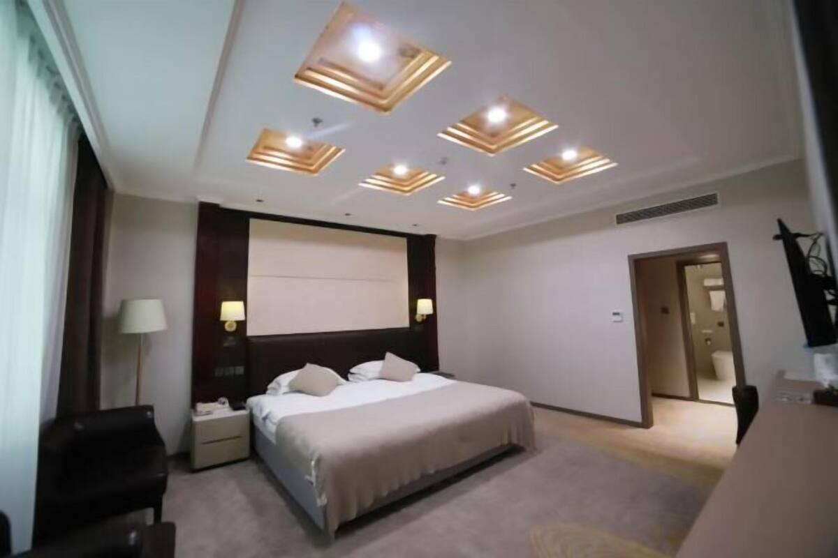 A hotel room with a bed, bedside tables, a desk, and a ceiling with recessed lighting.