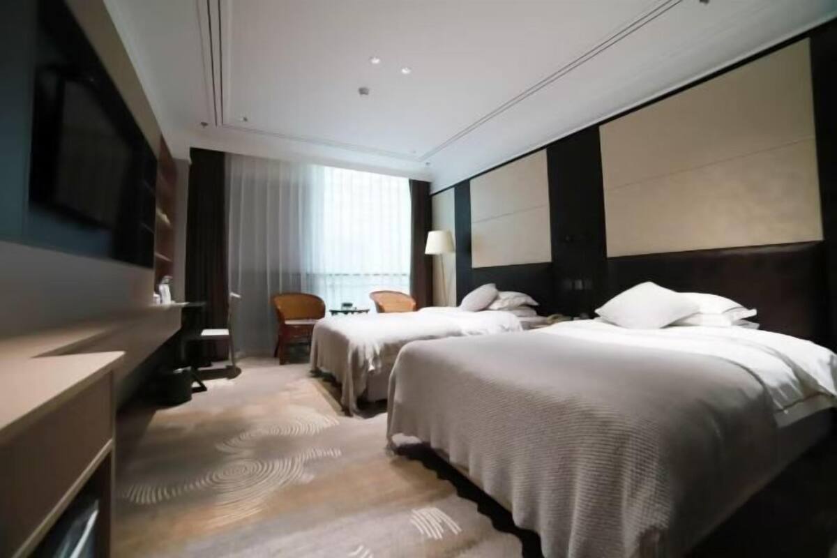 A hotel room with two beds, a desk, a chair, a lamp, and a large window.