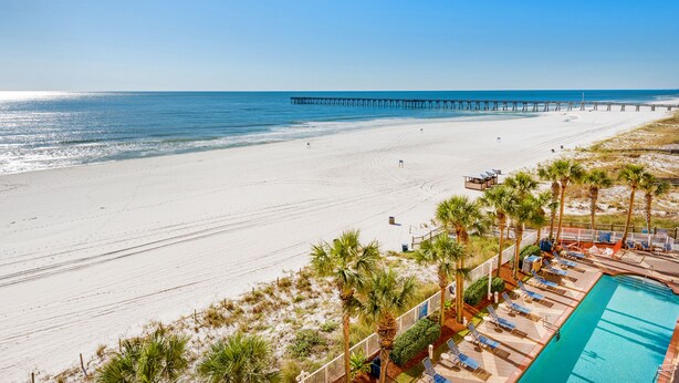 A beach view with a long pier extending into the ocean, a swimming pool with lounge chairs, and palm trees.