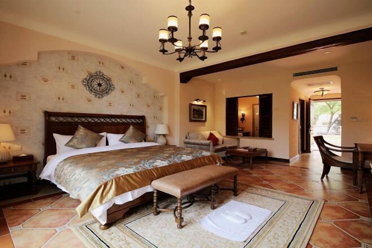 Een slaapkamer met een groot bed, een bankje, een kroonluchter, een behang met patroon en uitzicht op het omliggende landschap.