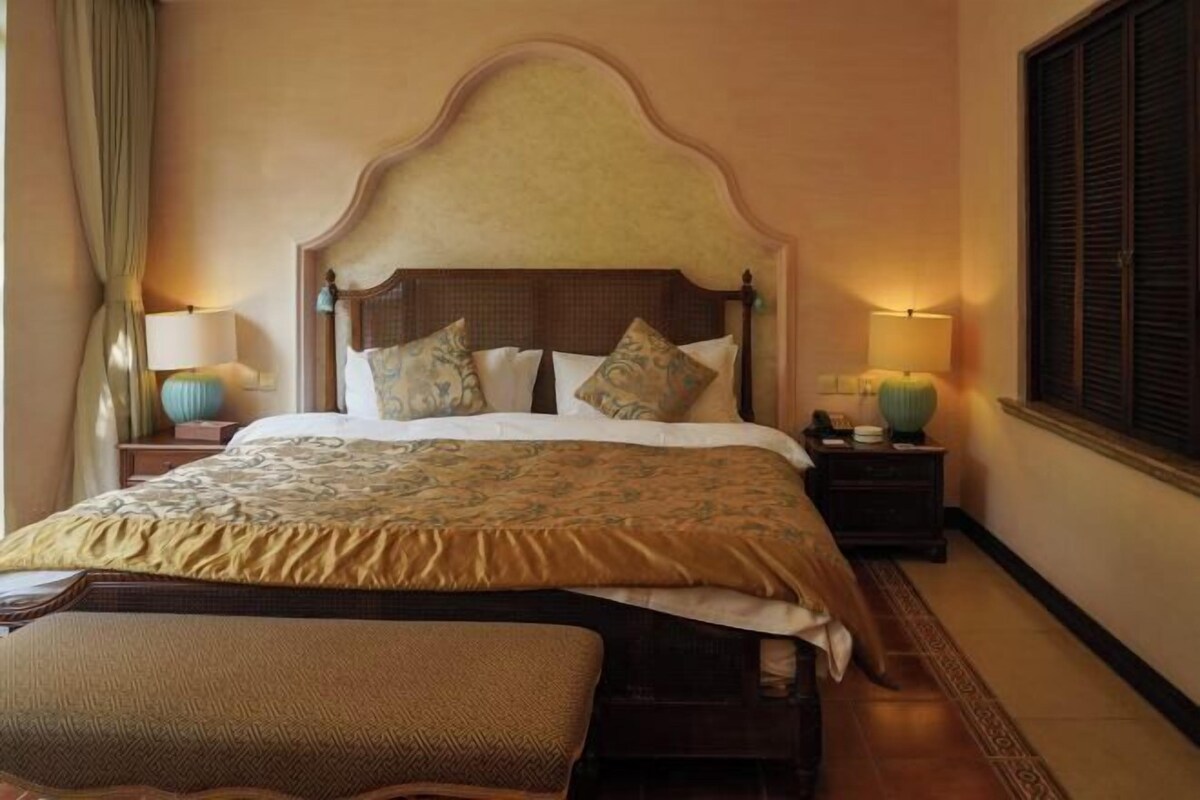 A hotel room with a large bed, two bedside tables with lamps, a bench, and a window with blinds.