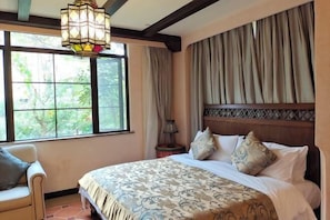 A bedroom with a large bed, a chair, a nightstand, a lamp, and a window with curtains.